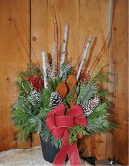 A black pot is filled with evergreen cuttings and white twigs. It is supplemented by red berries and pinecones. A large red ribbon, in a bow, is adhered to the front.