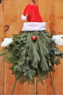 Evergreen tree cuttings are made to look like a gnome by being bunched together to make a triangle with a red santa hat, a red bell as a nose, and mittens on each side.