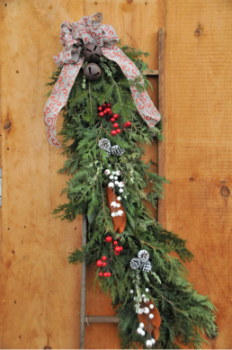 A wooden ladder is decorated with evergreen cuttings, red & white berries, and pinecones. It is topped by a red and grey ribbon, in a bow, with two bells.