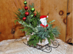 A small wooden sleigh is filled with evergreen cuttings, red bells, and green bells. A stuffed Sant Claus is placed at the front.