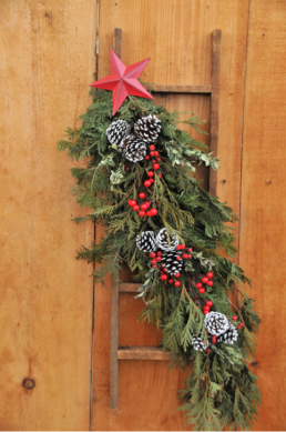 A wooden ladder is decorated with evergreen cuttings, red berries, and pinecones. It is topped by a red star.