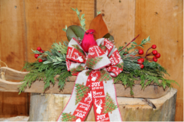 A small wooden sleigh is filled with evergreen cuttings and red berries. In the centre is a grey and red ribbon with a green Christmas tree and the words “JOY,” “HO HO HO,” and “Merry Christmas” is in a bow and topped by a red bird.