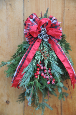 Evergreen cuttings are bunched together in a triangle and decorated with red berries. A black and red ribbon, in a bow, with three pinecones in the centre is placed on top.