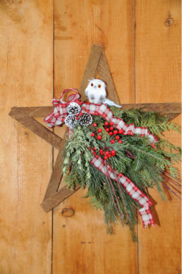 A wooden star is decorate in its centre with evergreen cuttings, red berries, a white and red ribbon, in a bow, and three pinecones. A white own sits at the top centre.