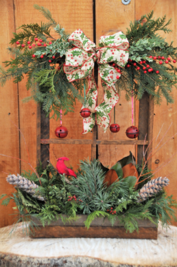 A wooden window frame. The window box at the bottom is filled with evergreen cuttings, pine cones, red berries, and a red bird. The window frame is topped with evergreen cuttings and red berries with a red, green, and white ribbon, in a bow, at the centre. Red bells also hang down i nthe middle of the window.